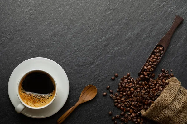 Top view above of Black hot coffee for morning with milk foam for morning menu in white ceramic cup with coffee beans roasted in sack bag on dark stone table background. Flat lay with copy space.