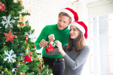 Mutlu Noeller ve yeni yıl konsepti. Noel temalı kıyafetleri olan genç beyaz bir sevgili Noel ağacını süslemeyi kutluyor ve evdeki partide kahkahalarla gülüp eğleniyor.