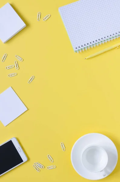 Top view office desk with notepads, phone, paper for notes, coffee cup on yellow background.