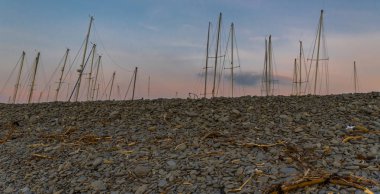 Yelkenli tekne direkleri, gün batımında, kırmızı gökyüzü karşı sürüklenen mal ve müsveddeleriyle ile çevrili bir çakıl plajı gösterilen