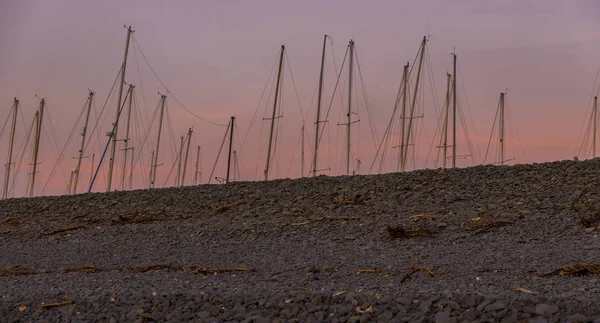 Navegando mastros de barco, mostrando atrás de uma praia de seixos, repleta de flotsam e jetsam, ao pôr do sol, contra um céu vermelho — Fotografia de Stock