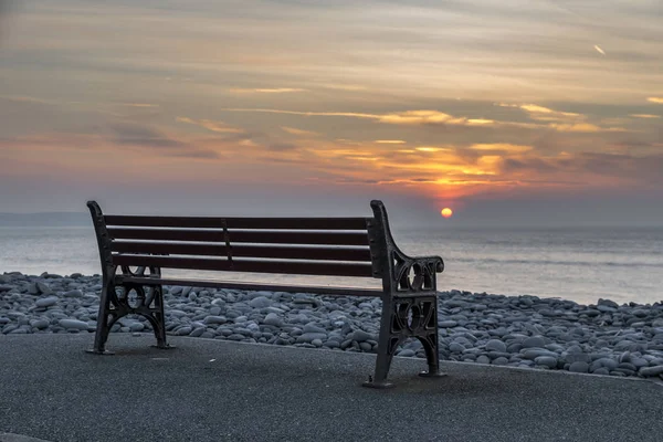 Un banco vacío, en la orilla del mar, con una puesta de sol ardiente y colorida en el océano — Foto de Stock