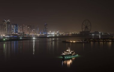 Geceleri Dubai Marina 'nın gökdelenleri. Modern binaların ışıkları, gece için demirlemiş lüks bir yatın bulunduğu binaların önündeki Basra Körfezi denizine yansıyor.