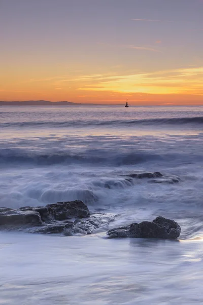 Waves rolling over rocks on the shoreline, while the sun sets over the horizon