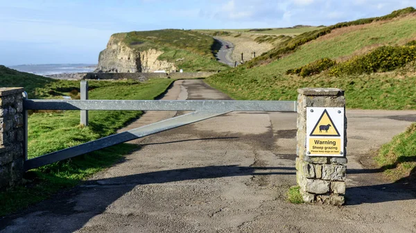 A closed gate, between two closed pillars, leading to a dramatic coast line. The pillar has a sign saying, \