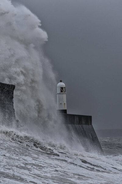 Huge waves breaking against a stone jetty, with a single lighthouse at the end, during a major winter storm. The location is Porthcawl, on the South Wales coast.