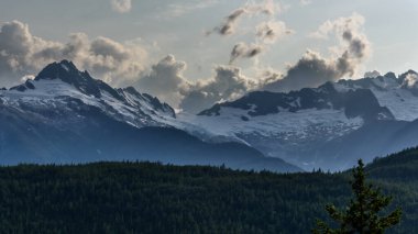 Dağlık bir ormanın karşısındaki karlı dağ tepelerinin görüntüsü ilginç bir gökyüzüne doğru ilerliyor. Dağlar Whistler 'a yakın, British Columbia, Kanada