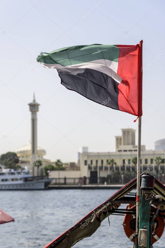 The UAE flag, flying the breeze, on Dubai Creek with the old district of Dubai, Bur Dubai, and a mosque minaret, in the background