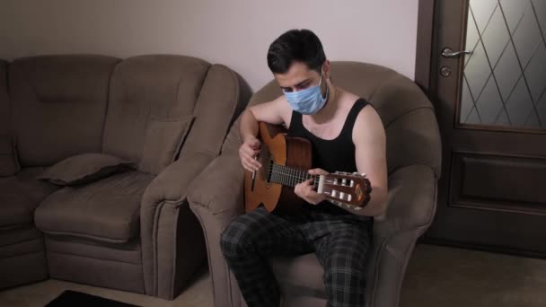 의료용 마스크를 쓴 백인 남성은 소파에 앉아 자립심을 가진 쿼런틴 (Quarantine) 에서 기타를 연주 한다. 기타를 연주하는 청소년 — 비디오