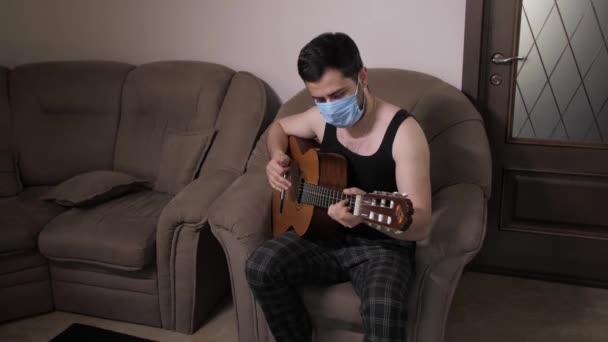 Tıbbi maske takmış beyaz bir adam kanepede oturmuş gitar çalıyor. Kendini tecrit karantinasında. Gitar çalan genç adam. — Stok video