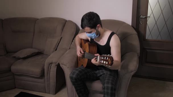 Homme caucasien dans un masque médical joue de la guitare assis sur le canapé auto-isolement quarantaine à la maison. Jeune homme jouant de la guitare — Video