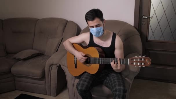 Blanke man met een medisch masker speelt gitaar zittend op de bank zelfisolatie quarantaine. Jonge man die gitaar speelt — Stockvideo