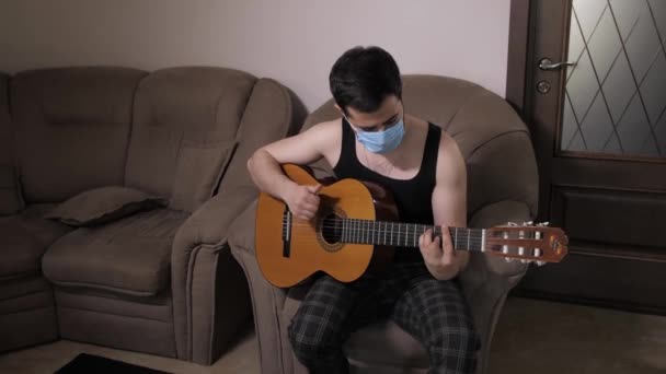 Blanke man met een medisch masker speelt gitaar zittend op de bank zelfisolatie quarantaine. Jonge man die gitaar speelt — Stockvideo