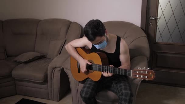 L'uomo caucasico con una maschera medica suona la chitarra seduto sul divano casa auto-isolamento Quarantena. Giovane ragazzo suonare la chitarra — Video Stock