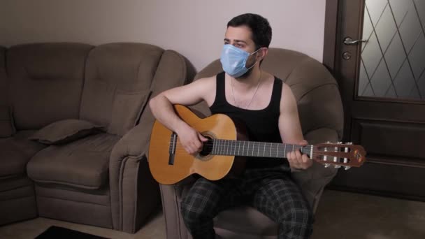 Biały mężczyzna w masce medycznej gra na gitarze, siedząc na kanapie i kwarantannie. Młody facet gra na gitarze — Wideo stockowe