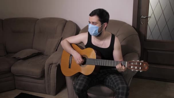 Белый мужчина в медицинской маске играет на гитаре, сидя на диване дома на карантине самоизоляции. Молодой парень играет на гитаре — стоковое видео