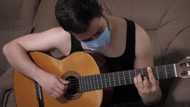 Белый мужчина в медицинской маске играет на гитаре, сидя на диване дома на карантине самоизоляции. Молодой парень играет на гитаре — стоковое видео
