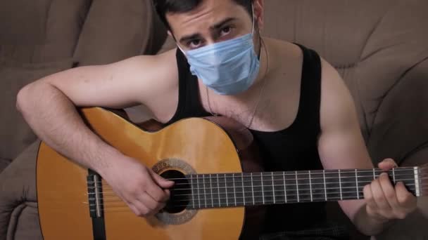 Blanke man met een medisch masker speelt gitaar zittend op de bank zelfisolatie quarantaine. Jonge man die gitaar speelt — Stockvideo