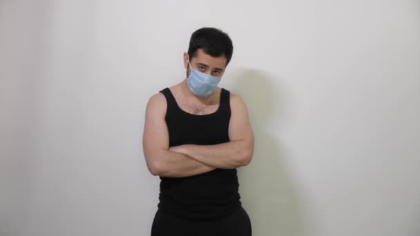 Um jovem caucasiano com uma máscara médica cruza as mãos na frente dele e olha para o fundo branco da câmera Covid19 Coronavirus — Vídeo de Stock