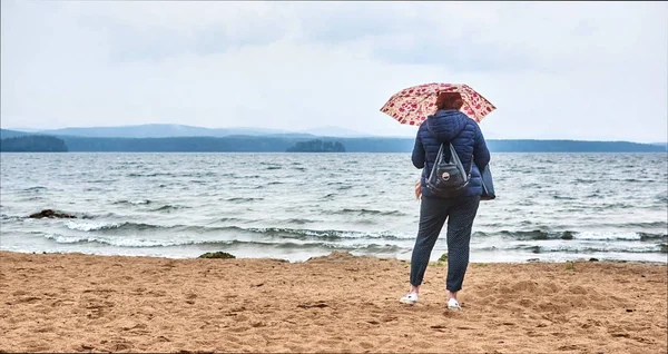 Şemsiyeli kızıl saçlı yalnız bir kız gölün kıyısında, kumların üzerinde duruyor. Orman. Dağlar. Güzel doğal manzara. Bulutlu gökyüzü. Doğada. Yaz günü. Yalnız genç kadın..