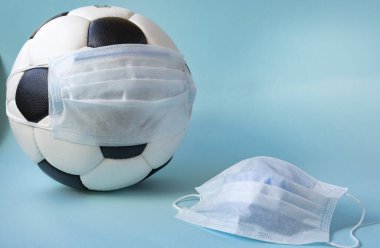 Mavi arka planda tıbbi maske takmış futbol topu. Virüslere karşı korona koruması bakterileri durdurur. Spor etkinliklerinin iptali. Boşluğu kopyala