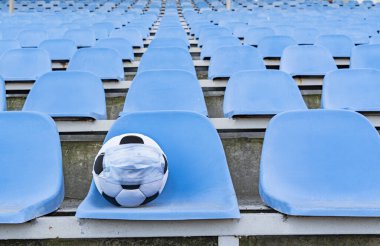 Boş stantların arkasında tıbbi maske takmış futbol topu. Tacın virüslerden ve bakterilerden korunması sona erer. Spor etkinliklerinin iptali. Açık bir stadyumda sıra sıra sandalye. Boşluğun bir kopyası.