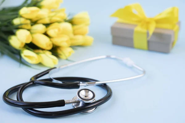 A bouquet of yellow tulips with a stethoscope and a gift on a blue background. National doctor\'s day. Happy nurse \'s day. Health day. Top view, a copy of the space.