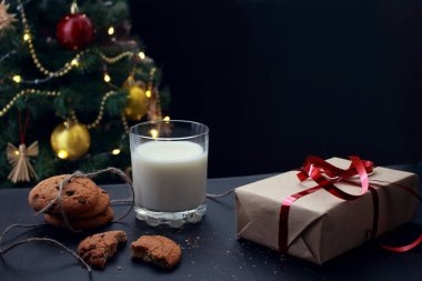 Bir bardak süt, çikolatalı kurabiyeler ve süslü bir Noel ağacının arka planında kırıntılar.