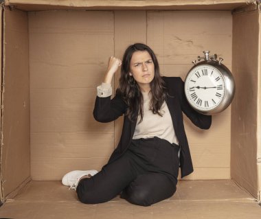 İş kadını elinde saatle karton bir ofiste oturuyor.