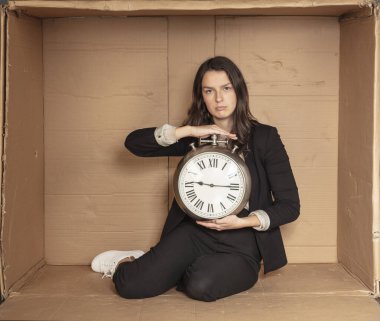 İş kadını elinde saatle karton bir ofiste oturuyor.