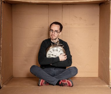 Genç işsiz bir öğrenci sıkı bir karton kutunun içinde oturuyor, deneyimsiz işsizlik fikri, zamanın geçişini simgeleyen bir saatin elindeki saat.