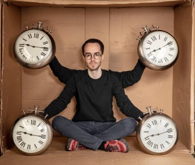 Genç işsiz bir öğrenci sıkı bir karton kutunun içinde oturuyor, deneyimsiz işsizlik fikri, zamanın geçişini simgeleyen bir saatin elindeki saat.