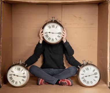 Genç işsiz bir öğrenci sıkı bir karton kutunun içinde oturuyor, deneyimsiz işsizlik fikri, zamanın geçişini simgeleyen bir saatin elindeki saat.