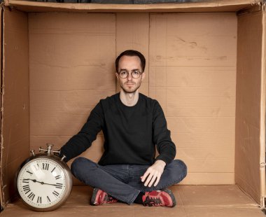 Genç işsiz bir öğrenci sıkı bir karton kutuda oturuyor, deneyimsiz işsizlik fikri, eldeki saat zamanın bir sembolü.