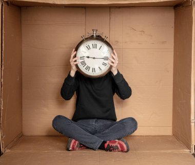 Genç işsiz bir öğrenci sıkı bir karton kutuda oturuyor, deneyimsiz işsizlik fikri, eldeki saat zamanın bir sembolü.