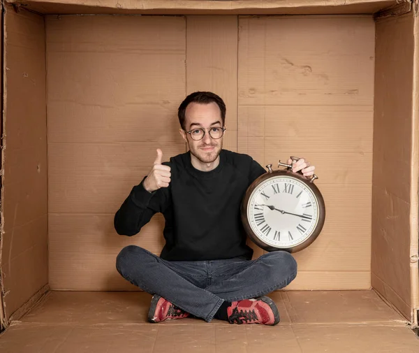 Genç işsiz bir öğrenci sıkı bir karton kutunun içinde oturuyor, deneyimsiz işsizlik fikri, zamanın geçişini simgeleyen bir saatin elindeki saat.