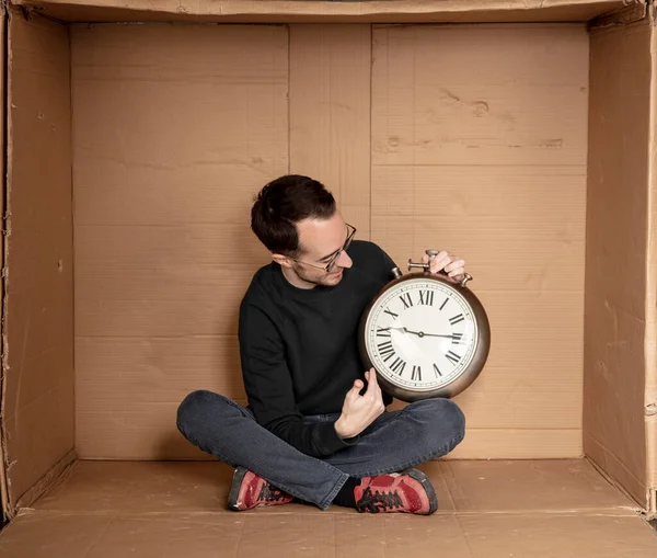 Genç işsiz bir öğrenci sıkı bir karton kutunun içinde oturuyor, deneyimsiz işsizlik fikri, zamanın geçişini simgeleyen bir saatin elindeki saat.