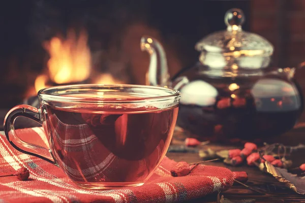 Tee mit Weißdorn in einer Glasschale und Teekanne auf einem Holztisch in einem Raum mit brennendem Kamin, Nahaufnahme — Stockfoto