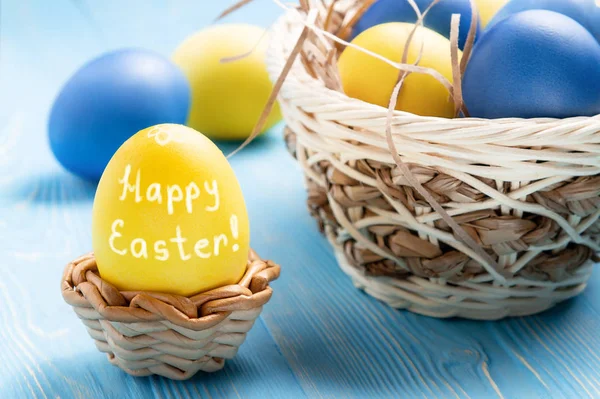 Easter greeting card - yellow egg in a wicker stand with the inscription Happy Easter and a basket with colored eggs on a blue wooden table