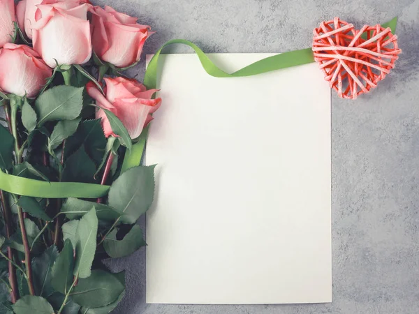 Arranjo de flor - um buquê de rosas rosa, um coração de vime e uma folha vazia da inscrição em uma superfície concreta, modelo do desenho ou cartão de saudação, visão superior, colocação chata — Fotografia de Stock