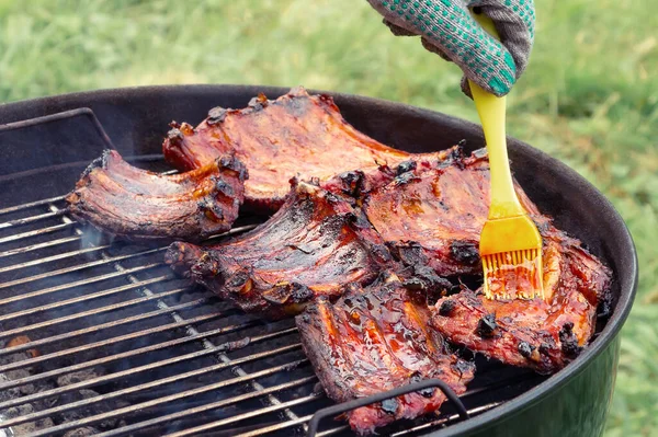 Grelhar costelas de porco em uma churrasqueira redonda, manchando com molho de marinada — Fotografia de Stock