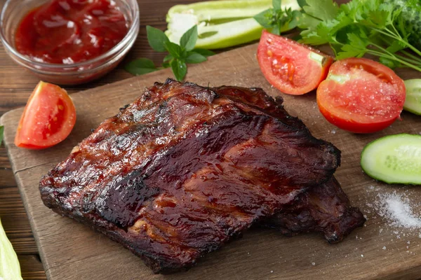 Costelas de porco churrasco em uma tábua de corte de madeira com ketchup e legumes — Fotografia de Stock