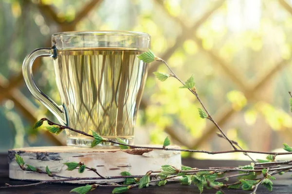 El jugo de abedul en una mesa en una taza de vidrio, junto a una rama de abedul con hojas jóvenes Imagen De Stock