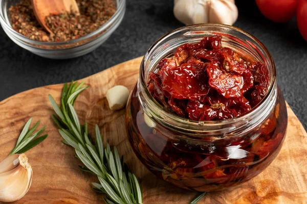 Sun-dried tomatoes with garlic, rosemary and spices in a glass jar on an olive wood cutting board — Stock Photo, Image