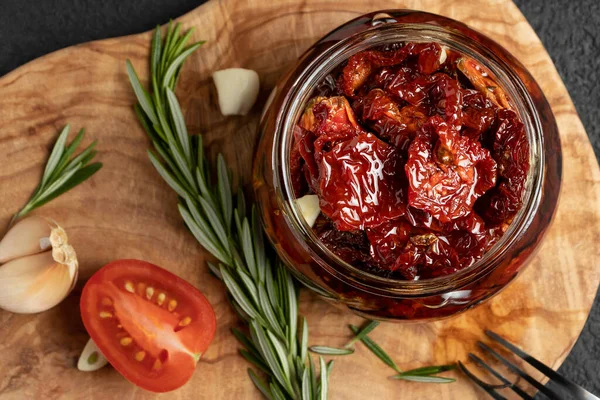 Tomates secados al sol con ajo, romero y especias en un frasco de vidrio sobre una tabla de cortar de madera de olivo, vista superior, puesta plana — Foto de Stock