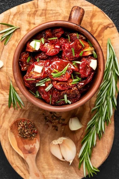 Tomates secados al sol con ajo, romero y especias en un tazón de arcilla sobre una tabla de cortar de madera de olivo, vista superior, puesta plana, imagen vertical — Foto de Stock