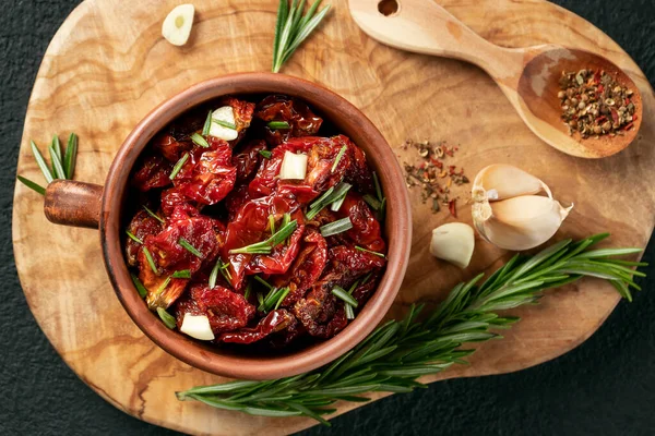 Tomates secados al sol con ajo, romero y especias en un tazón de barro sobre una tabla de cortar de madera de olivo, vista superior, puesta plana — Foto de Stock