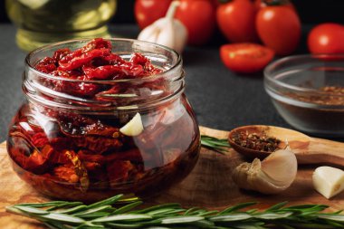 Sarımsaklı, biberiyeli ve zeytin ağacı kesme tahtasındaki cam kavanozda kurutulmuş domatesler.