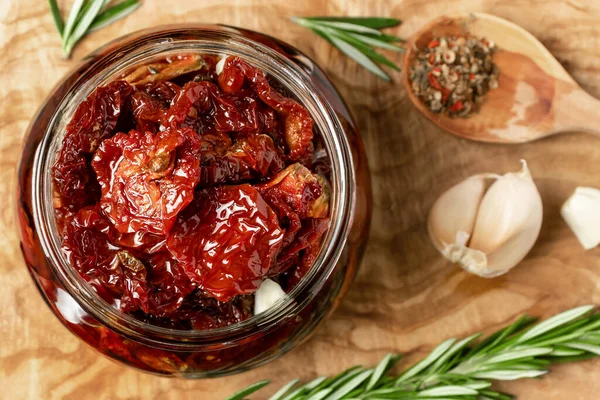 Sun-dried tomatoes with garlic, rosemary and spices in a glass jar on an olive wood cutting board, top view, flat lay — Stock Photo, Image