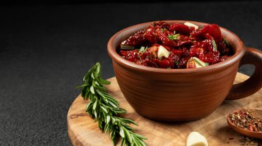 Sarımsaklı, biberiyeli ve baharatlı güneşte kurutulmuş domatesler zeytin ağacı kesme tahtasındaki kil kasesinde.